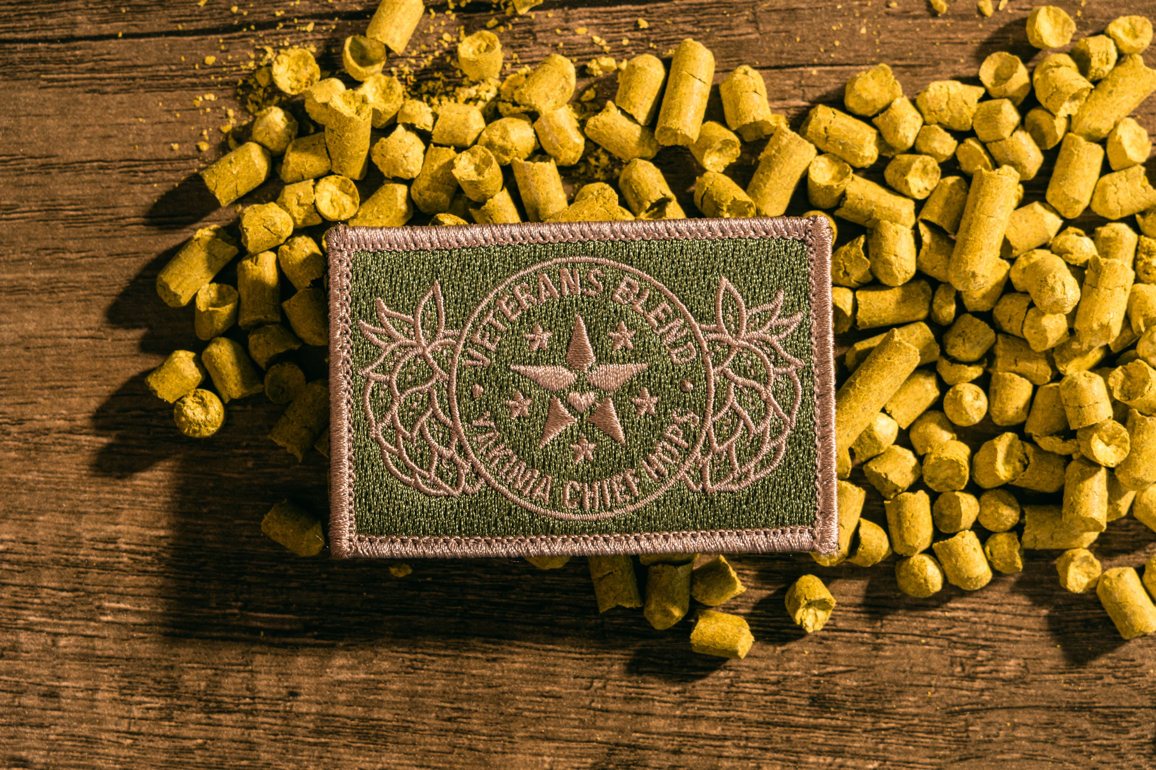 Military green patch with Veterans Blend emblem resting atop a pile of green hop pellets with wood backdrop