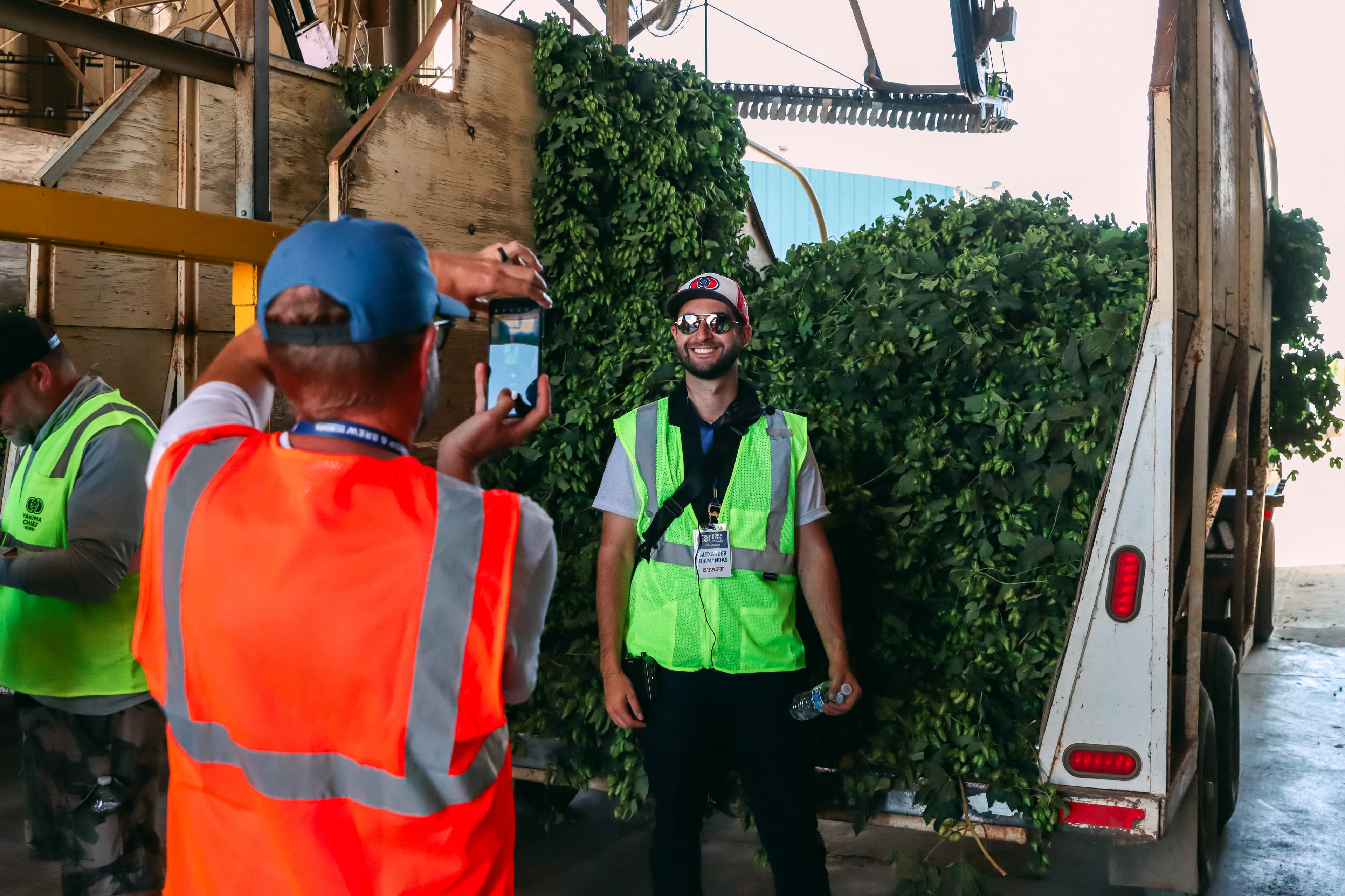Person in safety vest take picture on their phone of person standing in front of a hop truck filled with bines