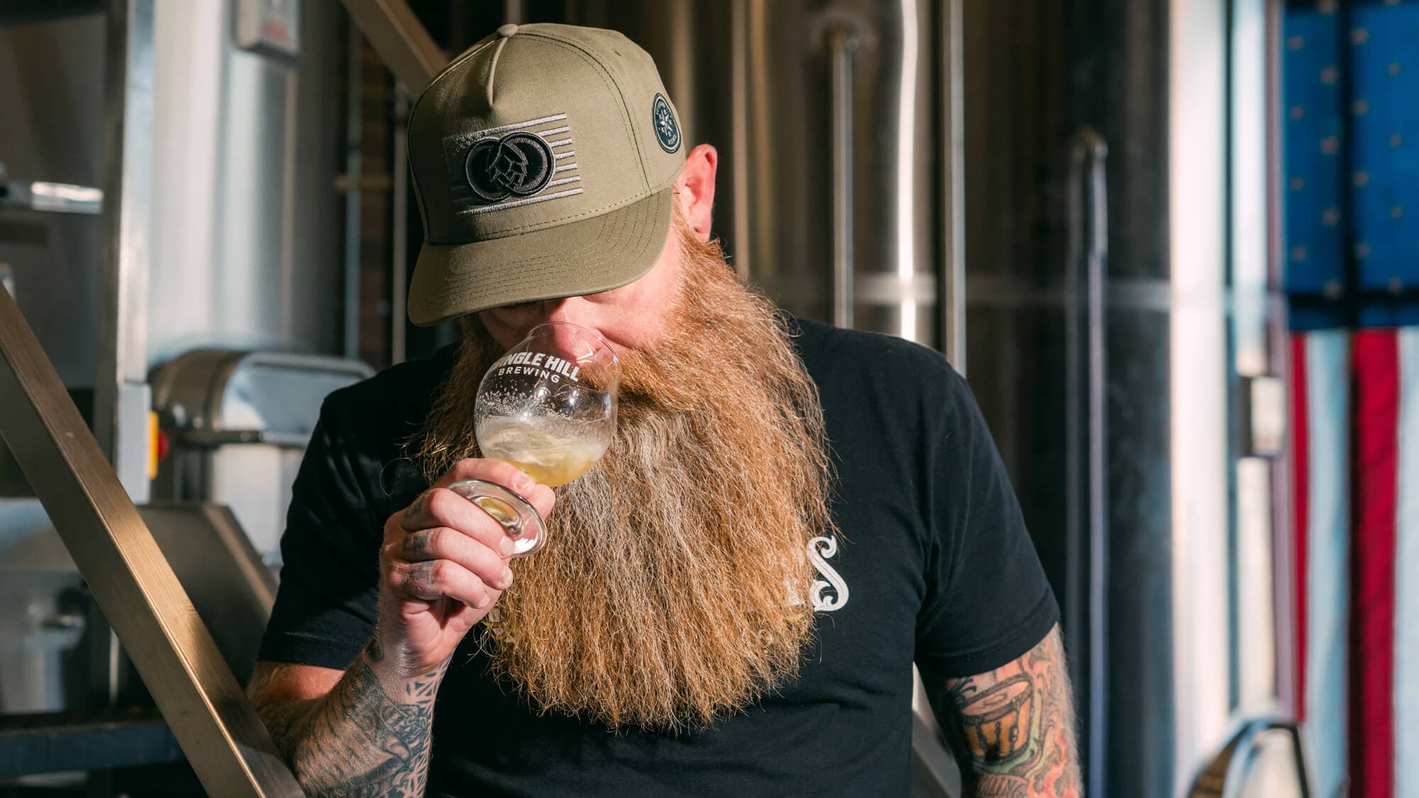 Bearded male brewer smelling beer in sample glass with American flag hanging in brewery as backdrop
