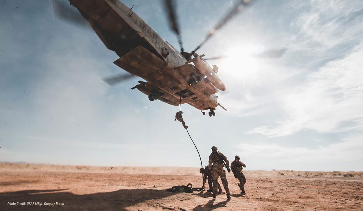 Soldiers on dusty ground as one rappels from a hovering helicopter