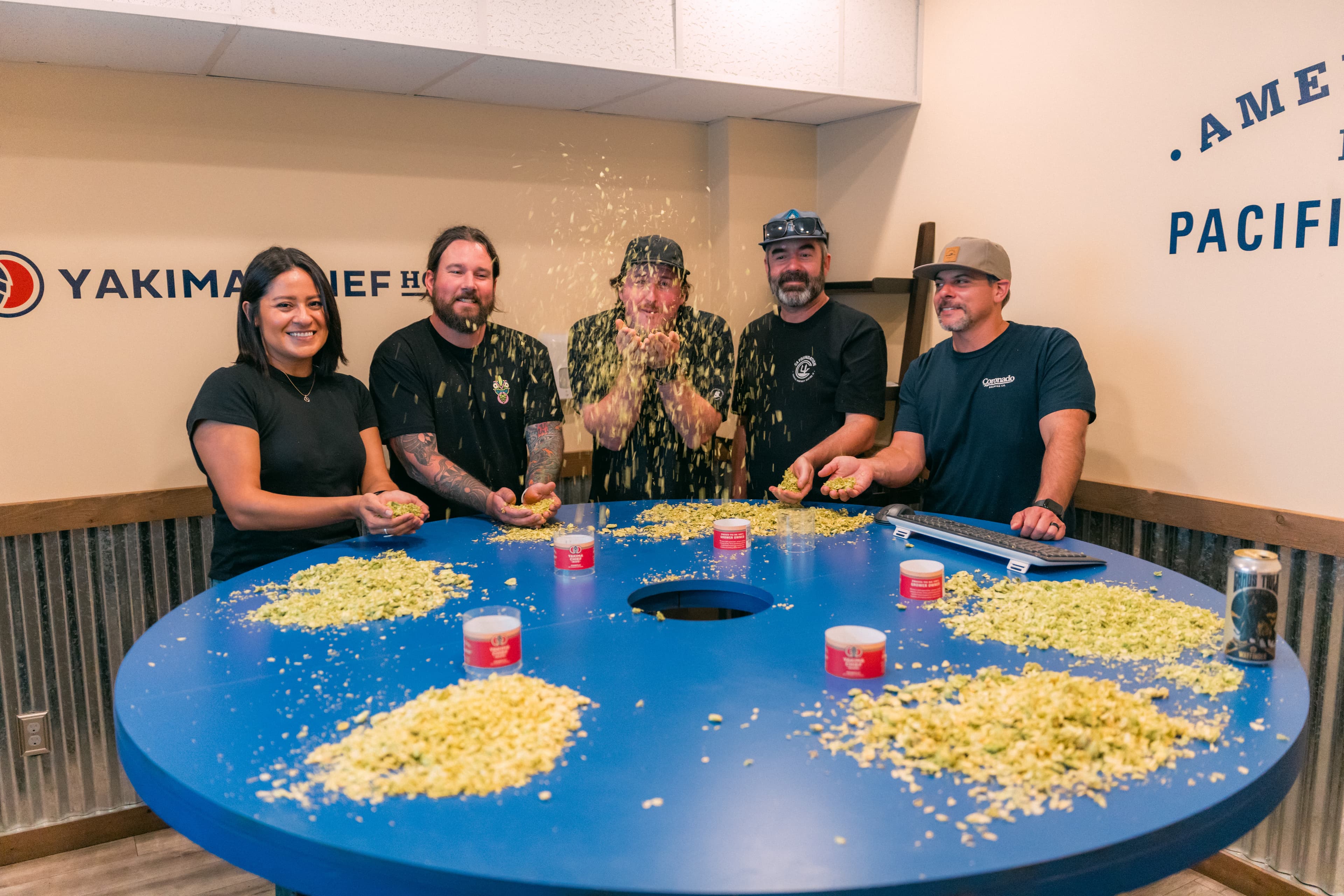 Brewers gathered in semi-circle around blue table covered with hops and tossing the leaves in air